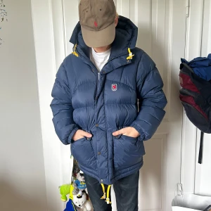 Blå dunjacka från Fjällräven med huva - Säljer en blå, pösig dunjacka från Fjällräven med huva och gula snören. Jackan har dragkedja och knappar framtill, två stora fickor och klassisk Fjällräven-logga på bröstet. Perfekt för kalla dagar när du vill hålla stilen och värmen. Skick 9/10! Som nyskick!