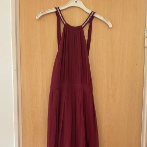 Vinröd långklänning med strassdetaljer - Vinröd långklänning från Nelly med plisserad överdel och smala axelband dekorerade med strass. Klänningen har en halterneck-modell och ett flowy fall. Perfekt till bal! Endast testad, nypris ca 1000kr.