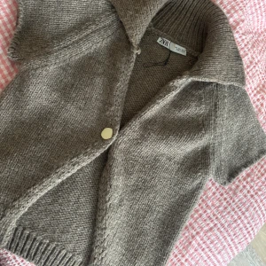 Beige stickad kofta från Zara - Kortärmad stickad kofta från Zara i beige nyans. Koftan har en stor krage, en dekorativ guldig knapp framtill och är gjord i ett mjukt, grovstickat material. Perfekt att slänga över ett linne för en chill look.