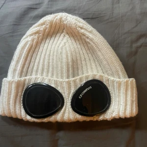 Mössa från C.P. Company - Vit ribbstickad mössa från C.P. Company med ikoniska svarta goggle-linser framtill. Mössan är tillverkad i mjuk ull och har en uppvikt kant. Perfekt för dig som vill sticka ut med en streetwear-inspirerad look. Den är lagad som ni ser på den andra bilden.