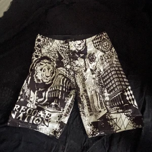 Reggae Nation shorts med lejonmotiv - Svarta och vita shorts från Reggae Nation med grafiskt tryck av lejon, byggnader och text. Snygg streetstil med raka ben och midja med knapp och dragkedja. Perfekta för dig som vill sticka ut med ett unikt mönster och coola detaljer.