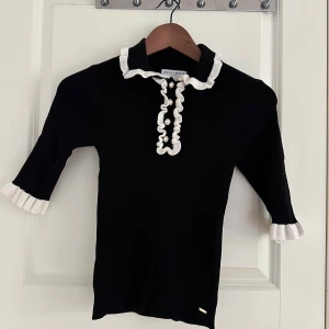 Svart pikétröja med volang - Supersöt svart pikétröja från Holly & Whyte med vita volangdetaljer vid krage, ärmslut och knäppning. Tröjan har pärlknappar framtill och trekvartsärm. 🖤