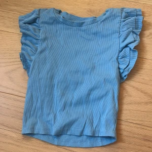 Blå ribbad topp med volangärm Zara - Supersöt blå ribbad topp från Zara med volangdetaljer på axlarna. Toppen är ärmlös och har en rund halsringning. Perfekt för dig som gillar en feminin och trendig stil.