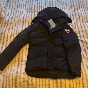 Svart dunjacka från Canada Goose  - Säljer en svart dunjacka från Canada Goose i storlek M. Jackan har huva, dragkedja och klassisk röd logotyp på ärmen. Perfekt för kalla dagar med sin fluffiga och varma design. Snygg och stilren modell som funkar till det mesta. 