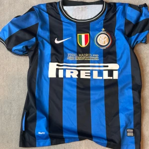 Inter Ronaldo 9 fotbollströja Nike - Säljer en klassisk Inter fotbollströja med Ronaldo 9 på ryggen. Tröjan är blå och svart med breda ränder, officiella klubbmärken, Pirelli-tryck och Champions League-märke på ärmen. Tillverkad i lätt och ventilerande polyester.