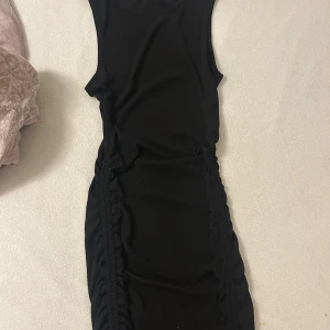 Svart tight ärmlös kortklänning - Svart tight ärmlös klänning med rynkade sidor som ger en snygg siluett. Modellen är kort och har en enkel, stilren design utan synliga detaljer framtill. Perfekt för dig som gillar minimalistisk och trendig stil.