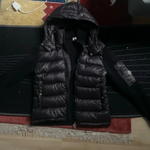 Svart Moncler cardigan med huva - Snygg svart cardigan från Moncler med stickade ärmar och vadderad kropp. Jackan har huva, dragkedja framtill och en ikonisk Moncler-logga på ärmen. Perfekt för kyliga dagar och ger en riktigt clean look.