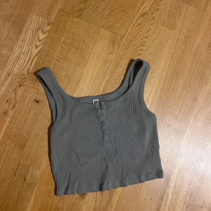 Grön ribbad linnetopp från Uniqlo - Säljer en grå ribbad linnetopp från Uniqlo med croppad passform. Toppen har breda axelband och knappar framtill som ger en snygg detalj. Perfekt att matcha med jeans eller kjol för en chill look.