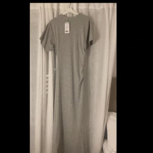 Grå långklänning med kort ärm - Helt oanvänd med prislapp kvar. Enkel och stilren grå långklänning i mjukt material. Klänningen har rund halsringning och korta ärmar, tajtare i midjan, perfekt för en avslappnad och trendig look. Passar dig som gillar minimalistisk stil och vill ha något bekvämt men snyggt. 