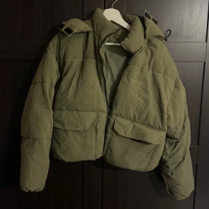 Olivgrön pufferjacka XS - Säljer en olivgrön pufferjacka i storlek XS. Jackan har en croppad passform, stora fickor framtill och dragkedja. Perfekt för kalla dagar med sin vadderade look och höga krage. Snygg och trendig modell som håller dig varm.