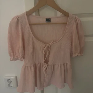 Ljusrosa blus  - Supersöt ljusrosa blus från Gina tricot med puffiga korta ärmar och två knytningar framtill. Blusen har en lätt rynkad struktur och volang nedtill, perfekt för en romantisk look. Djup urringning och luftig passform.