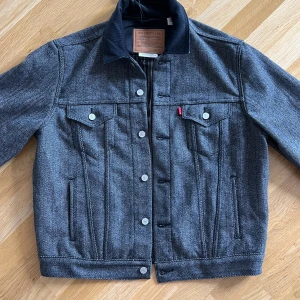 Levi's Premium Vintage Truckerjacka M - Snygg vintageinspirerad truckerjacka från Levi's Premium i grått fiskbensmönster. Jackan har klassisk jeansjackeform, knappar framtill, två bröstfickor med lock och en svart krage i kontrastmaterial. Perfekt för dig som gillar retrovibbar och streetstyle.