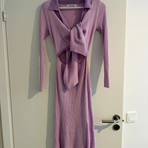 Lila ribbad långklänning från Adoore - Superfin lila långklänning från Adoore med ribbad struktur, lång ärm och snygg krage. Klänningen har en stor knyt-detalj framtill och cut out vid midjan som ger en trendig look. Perfekt för dig som vill sticka ut med en cool och modern stil.