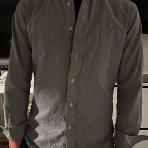 Grå skjorta från Ralph Lauren - Klassisk grå skjorta från Ralph Lauren i slim fitt modell med button-down krage och broderad logga på bröstet. Skjortan har lång ärm, knappar framtill och är tillverkad i mjuk bomull. Perfekt för en clean och stilren look.