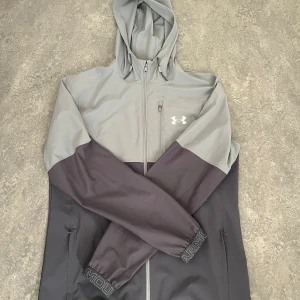 Grå och svart vindjacka från Under Armour - Snygg tvåfärgad vindjacka från Under Armour i grått och svart. Jackan har huva, dragkedja framtill, bröstficka med dragkedja och logga på bröstet. Ärmarna har elastiska muddar med broderad text. Perfekt för blåsiga dagar. (Denna version säljs ej längre. Pris kan diskuteras vid snabb affär!