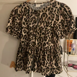 Leopardmönstrad blus med puffärm - Säljer en beige och svart leopardmönstrad blus med puffiga korta ärmar och två knytningar framtill. Blusen har rund halsringning och volang nedtill för en extra söt look. Jätte bra skick, som ny