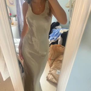 Champagnefärgad långklänning i satin - Säljer en elegant långklänning i skimrande champagnefärgad satin. Klänningen har en figurnära passform, ärmlös design och djup, öppen rygg.  Aldrig använd från Asos