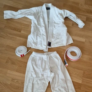 Vit judodräkt med två bälten - Judo-/kampsport dräkt i vitt från Budo Nord, komplett med jacka och byxor samt två bälten med röda markeringar från Jiu Jiutsu graderingar. Dräkten har klassisk omlottjacka med förstärkta sömmar och byxor med dragsko i midjan.