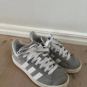 Grå Adidas Campus sneakers i mocka - Säljer ett par grå Adidas Campus sneakers med klassiska vita ränder och vit sula. Skorna har ovandel i mjuk mocka och vita skosnören. Perfekt för dig som gillar en clean och stilren look med retrovibbar. Varan har små defekter och visar liten tecken på andvändning. Är bara andvänd ett fåtal gånger💗