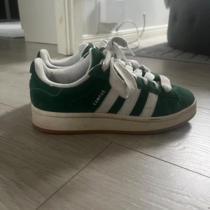 Adidas Campus gröna sneakers mocka - Gröna Adidas Campus sneakers i mocka med klassiska vita ränder och vit sula. Skorna har rund tå, platt sula och snörning. Adidas logga på hälen och Campus-tryck på sidan. Skorna är i bra skick, köpta förra våren för runt 1000kr. 