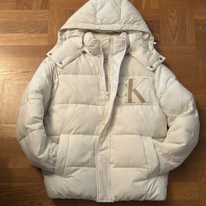 Vit pufferjacka från Calvin Klein Jeans - Snygg vit pufferjacka från Calvin Klein Jeans med stor CK-logga på bröstet. Jackan har huva med snörning och tryckknappar, dragkedja framtill och quiltad design. Perfekt för kalla dagar och riktigt clean look.