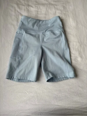 Ljusblå träningsshorts från MOVE - Säljer ett par ljusblå träningsshorts från MOVE med hög midja och praktiska sidofickor. Shortsen är gjorda i mjukt och stretchigt syntetmaterial som passar perfekt för gymmet eller löprundan. Snygg, enkel design med MOVE-logga på linningen.