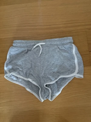 Grå mjuka shorts med vit kant - Grå shorts i mjukt material med vit kant och snörning i midjan. Sportig look med elastisk midja och kort passform, perfekta för varma dagar eller träning. Klassisk design som är enkel att matcha med olika toppar. 95% Bomull, 5% Viskos.