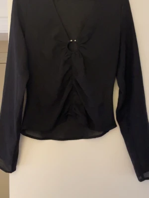 Svart omlottopp med ringdetalj Gina Tricot - Säljer en svart omlottopp från Gina Tricot i storlek M. Toppen har lång ärm, v-ringad hals och en snygg ringdetalj med pärlor framtill. Materialet är lätt och tunt, perfekt för att styla med jeans eller kjol. Passformen är normal och toppen har en croppad känsla.