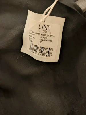Svart långklänning Line of Oslo XS - Svart långklänning från Line of Oslo i modellen Priscilla Solid. Klänningen är i ett tunt, transparent tyg med v-ringning och långa ärmar. Den har en luftig passform och faller snyggt ner mot anklarna. Perfekt för dig som gillar stilrena och enkla plagg.