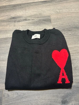 Svart stickad tröja från Ami - Svart stickad tröja från Ami med rund hals och ribbade muddar. På bröstet finns ett stort rött hjärta med bokstaven A under, vilket ger en unik och snygg detalj. Perfekt för dig som gillar stilrena plagg med en twist.