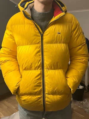 Gul pufferjacka från Tommy Jeans - Säljer en gul pufferjacka från Tommy Jeans med huva och dragkedja framtill. Jackan har ett broderat logomärke på bröstet och är fodrad för extra värme. Perfekt för kalla dagar och har en riktigt schysst streetstil.