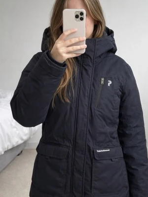 Peak Performance XS vinterjacka parkajacka - Svart parkajacka från Peak Performance i storlek XS. Jackan har huva, flera fickor med dragkedja och logga på bröstet och huvan. Materialet är slitstark polyester med dunfoder och meshdetaljer på insidan. Trasig mesh på innerfickorna, i övrigt i fint skick. Därav lågt pris. 