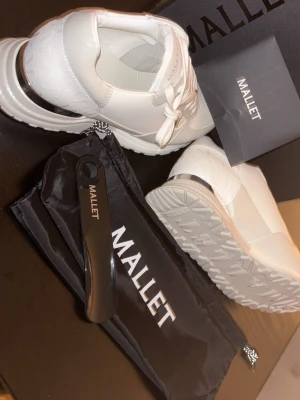 Vita sneakers från Mallet london  - Säljer ett par fräscha vita sneakers från Mallet med snygg metallic-detalj på hälen. Skorna har snörning, grov sula och är tillverkade i en mix av skinn och syntet. Perfekta för dig som vill ha en clean och modern look. Helt nya och inte använda , ny pris 2500 , säljs för 1950. 
