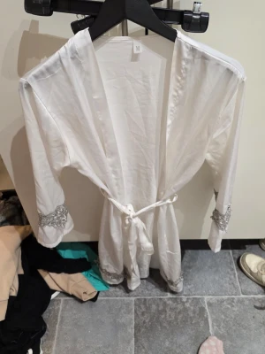 Vit kimono med spetsdetaljer - Säljer en vit kimono i mjukt tyg med knytband i midjan. Kimono har snygga spetsdetaljer i silver vid ärmsluten som ger en elegant touch. Perfekt för dig som vill ha något extra till din outfit.