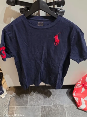 Mörkblå t-shirt från Polo Ralph Lauren - Snygg mörkblå t-shirt från Polo Ralph Lauren med rött broderat logomärke på bröstet och en stor röd siffra 3 på ärmen. Klassisk rund hals och korta ärmar. Tillverkad i mjuk bomull som känns skön mot huden. Perfekt för dig som gillar stilrena märkesplagg.