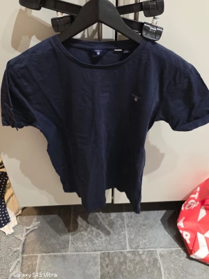 Mörkblå t-shirt från GANT - Snygg mörkblå t-shirt från GANT med klassisk rund hals och liten broderad logga på bröstet. T-shirten är kortärmad och gjord i mjuk bomull, perfekt för chill dagar. Enkel och stilren design som funkar till det mesta.