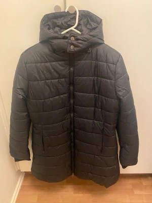 Svart pufferjacka med huva - Säljer en svart pufferjacka med huva och dragkedja framtill. Jackan har quiltad design och två fickor med dragkedja på sidorna. Perfekt för kalla dagar och har en stilren look som funkar till många outfits.