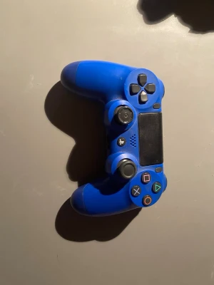 PlayStation DualShock 4 Wireless Controller - Blå PlayStation DualShock 4 trådlös handkontroll i gott skick. Alla knappar och styrspakar syns tydligt, och inga större repor eller skador är synliga. Perfekt för PlayStation 4-spelare som vill ha en extra eller ersättningskontroll. Lite trasig vänster spak.