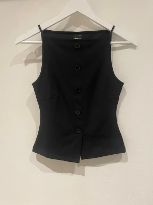 Svart knappad korsettopp från Gina Tricot - Säljer en svart korsettopp från Gina Tricot i storlek XS. Toppen har en figurnära passform, knappar framtill och smala axelband. Materialet är mjukt och känns som polyester. Perfekt att styla med jeans eller kjol för en cool look.