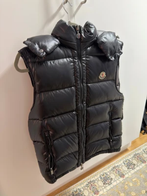 Svart dunväst från Moncler - Säljer en svart dunväst från Moncler med quiltad design och hög krage. Västen har två fickor med dragkedja och Moncler-logga på bröstet. Perfekt att slänga över en hoodie för extra värme. Stängs med dragkedja framtill.