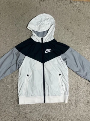 Nike windbreaker Youth storleken M - Nike vindjacka / träningsjacka – Youth M  Snygg och sportig Nike-jacka i färgkombinationen vitt, svart och grått. Perfekt som vindjacka eller lätt vår/höstjacka.  Redo att skickas snabbt.  OBS: Det finns en liten fläk på högra armen ( Kolla bild 5)