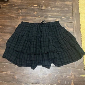 Svart rutig volangkjol med snörning - Kort svart kjol med rutigt mönster och flera volanger. Kjolen har en bekväm resår i midjan med snörning för justerbar passform. Perfekt för dig som gillar en avslappnad men ändå trendig stil.