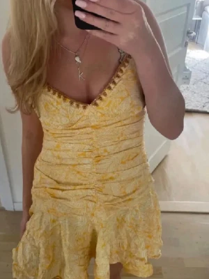 Gul kortklänning med blommönster Zara - Supersöt gul kortklänning från Zara med tunna axelband och v-ringning dekorerad med små broderade detaljer. Klänningen har ett blommigt mönster, rynkade partier framtill och en volangig kjol. Perfekt för soliga dagar och sommarhäng!
