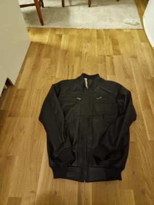 Svart bomberjacka i skinnimitation - Säljer en svart bomberjacka från EM med skön krage och muddar vid ärmslut och midja. Jackan har dragkedja framtill, två bröstfickor med dragkedja och två sidofickor med dragkedja. Materialet är skinnimitation och insidan är fodrad med mjukt tyg.