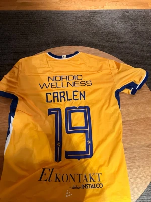 IFK Göteborg matchtröja gul XL - Galet snygg gul MATCHANVÄND matchtröja av Adam Carlén, från Ifk Göteborg och Craft med blå detaljer och tryck. Tröjan har klubbmärke, sponsorloggor och nummer 19 med namnet Carlén på ryggen. Materialet är lätt och andas, perfekt för fotboll. Snygg design med blå och vita ränder på sidorna. Kom gärna med prisförslag och hör av dig minsta lilla fundering, jag svarar alltid så fort jag kan 🤩