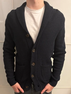 Mörkblå stickad kofta med knappar - Säljer en mörkblå stickad kofta med grov struktur och stora bruna knappar framtill. Koftan har två fickor, bred krage och lång ärm. Perfekt för lager på lager under kyliga dagar.