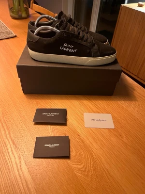 Svarta Saint Laurent sneakers - Svarta sneakers från Saint Laurent med vit broderad logga på sidan och vit sula. Skorna har snörning och är tillverkade i canvas. Klassisk låg modell med rund tå och diskreta detaljer. Perfekta för dig som gillar stilrena och exklusiva sneakers. Desvärre inte autentiska men extremt lika och otroligt bra kvalité ändå! Hör av dig vid minsta fundering och tvivla inte på att lägga ett prisförslag!🤩