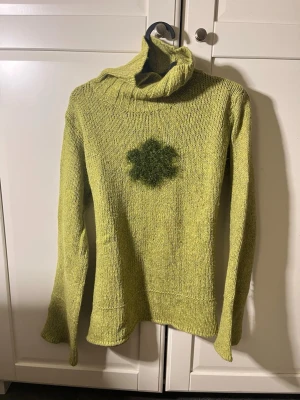 Grön stickad polotröja med blomma - Säljer en grön stickad polotröja med hög krage och utsvängda ärmar. Tröjan har en mörkgrön blommig detalj mitt på bröstet som ger en unik look. Perfekt för dig som vill ha något annorlunda och mysigt i garderoben.