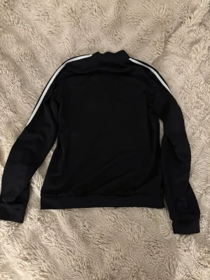 Svart Adidas zip-tröja med vita ränder - Säljer en svart Adidas zip-tröja med klassiska vita ränder längs ärmarna och Adidas-logga på bröstet. Tröjan har dragkedja framtill och två fickor med zip. Perfekt för en sportig och chill stil.