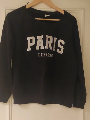 Svart sweatshirt Paris Gina Tricot - Cool svart sweatshirt från Gina Tricot med trycket 'PARIS LE MARAIS' i vitt framtill. Tröjan har rund hals, långa ärmar och en relaxed passform. Perfekt för dig som gillar streetstyle och vill ha en enkel men snygg look.